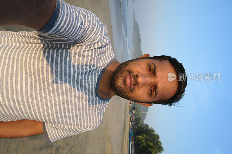 20岁出头的亚洲印度游客在海滩自拍，拿着以海滩为背景的手机在度假果阿海滩用手机、t恤、沙子、海边的日落/日出自拍